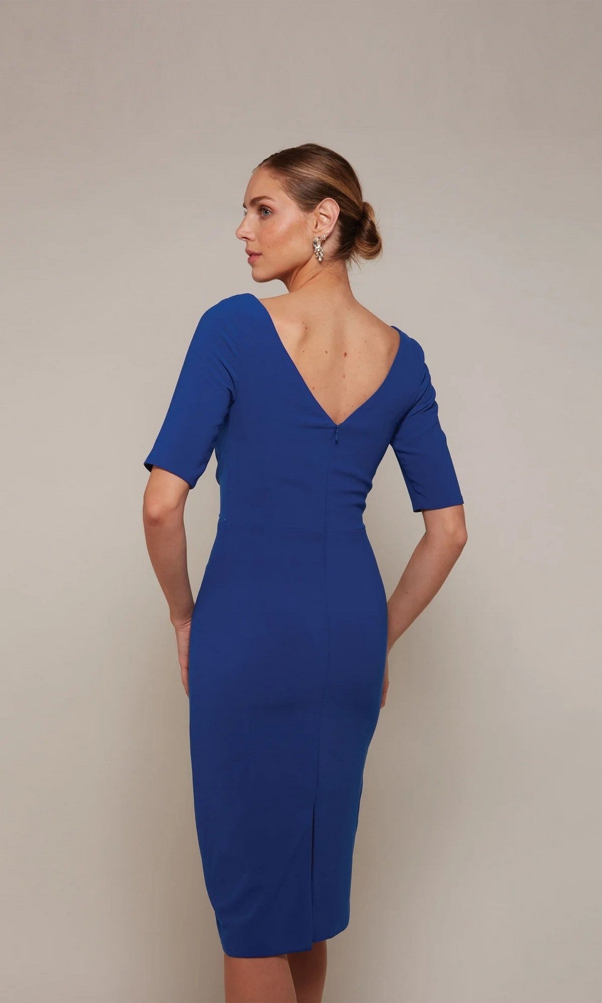 A woman wearing the Alyce 70036 Knee Length Semi-Formal Dress—a royal blue fitted dress with a V-back and elbow-length sleeves—stands against a neutral background.